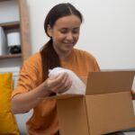 Happy young woman unboxing international package sitting on coach in living room of accommodation